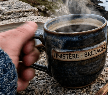 Coffee mug labeled Finistère-Bretagne overlooking rocky cliffs and ocean at sunset